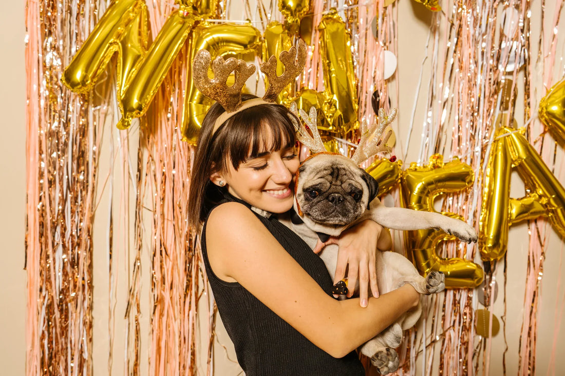 A brunette woman is hugging/holding a pug in her arms. They are both wearing headbands with antlers on them. There is a pink background with metallic streamers and gold "New Years" balloons. 